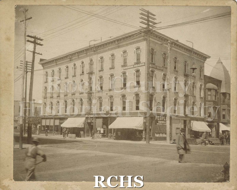 Black and white photograph of Business College building on corner of Main and Milwaukee. Sign for People's Drug Company visible on right corner.