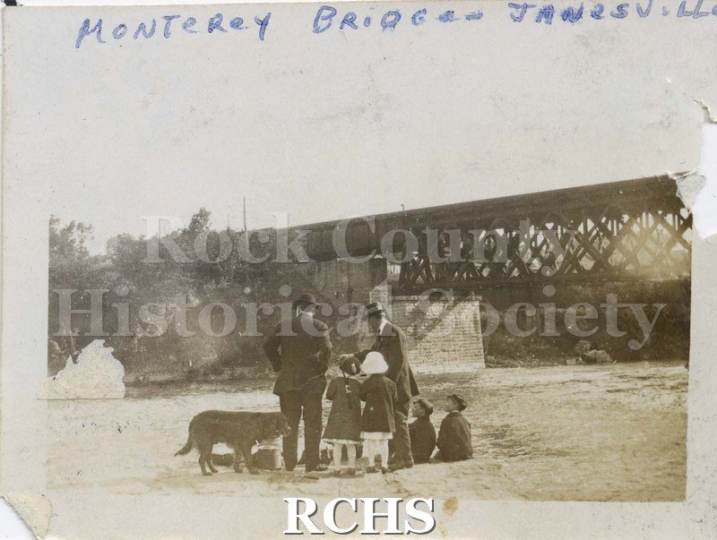 Black and white photograph of Monterey Bridge, Janesville, WI. Shows two unidentified men; four unidentified children and a dog standing below the bridge on the.