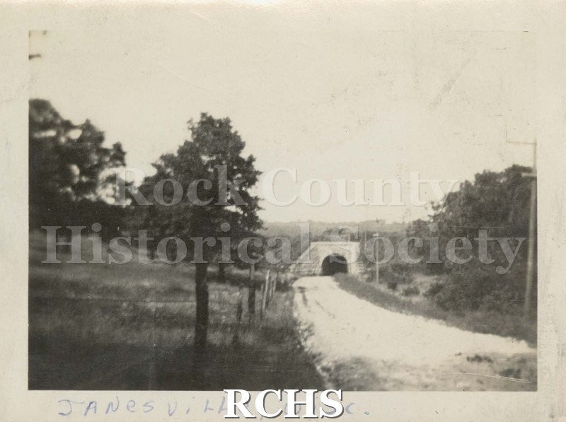 Black and white photograph of Janesville, WI, 1921 Tunnel. Shows unpaved road leading to a tunnel with trees on either side and power lines on right side.