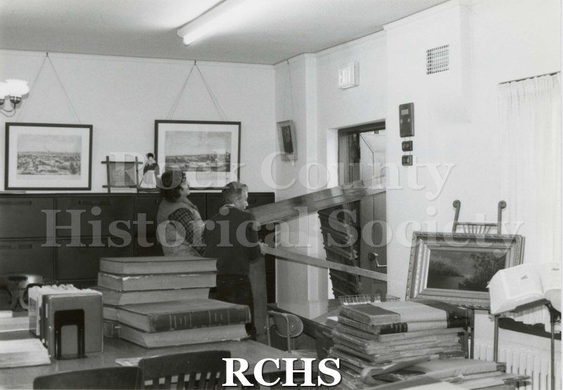 Black and white photograph of Beloit Newspapers. Shows inside of office. Shows two men moving in a piece of furniture. On the desks are stacks of books.