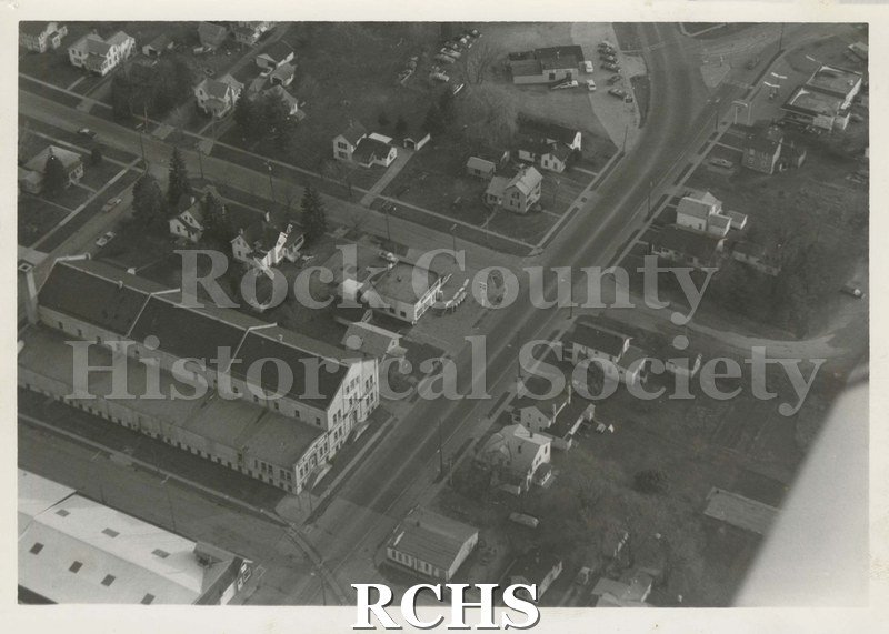 Black and white aerial photograph of Edgerton, WI. Shows a factory building on the left and houses to the right and left.