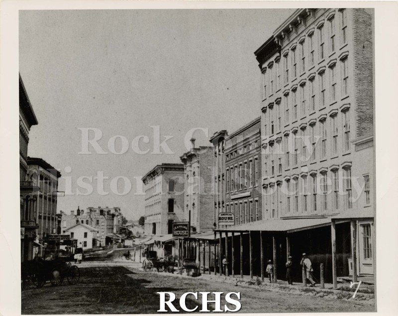 Black and white photograph of Janesville, WI, c. 1875, looking west from N. Bluff (now Parker) Street. Shows storefronts, especially American House.