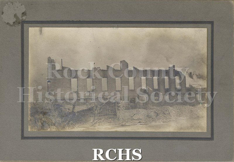 Postcard. Tinted image of the dam across the Rock River in Janesville. A young boy is standing on top of a stone wall on the edge of the dam with a fishing pole.