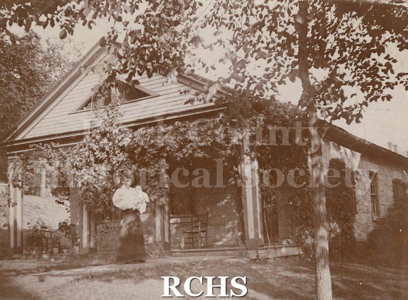 Black and white photograph of the Little Stone House. Shows a woman and child standing in front of it. There are vines covering the front.