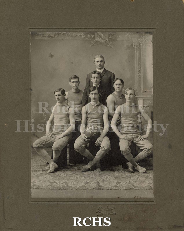 Black and white photograph of Janesville High School Basketball Team 1904-1905. There are six young men sitting with a basketball on which is printed "J.H.S. '05".