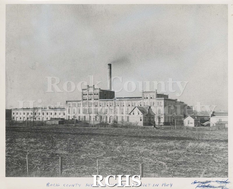 Black and white photograph of Rock County Sugar Co., showing the building in the middle and railcars in front. On bottom: "Rock County Sugar Co. Built in 1904".