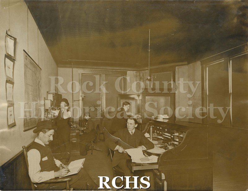 Tinted photo of Wisconsin Carriage Company workers. Four unidentified men and one unidentified woman are working in an office.