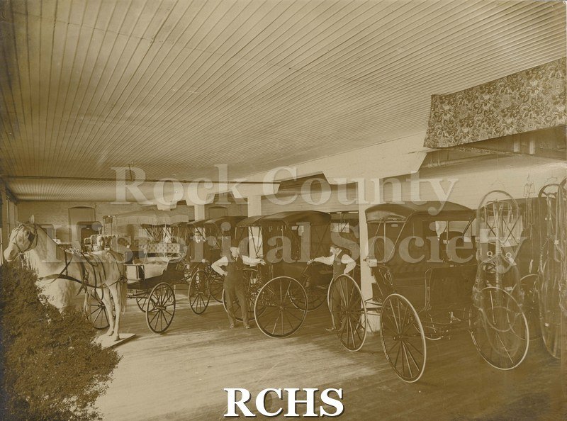 Black and white photograph of Wisconsin Carriage Company buggies on display. Three unidentified men are standing by finished buggies.