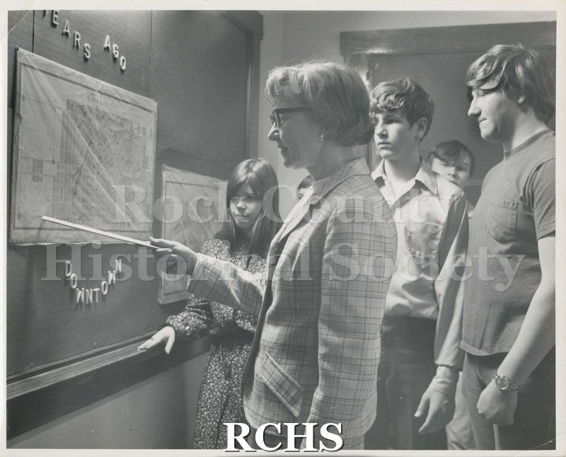 Black and white photograph of Marshall Junior High School Mini Course class. Students are standing around a teacher who is pointing to a map on the bulletin board.
