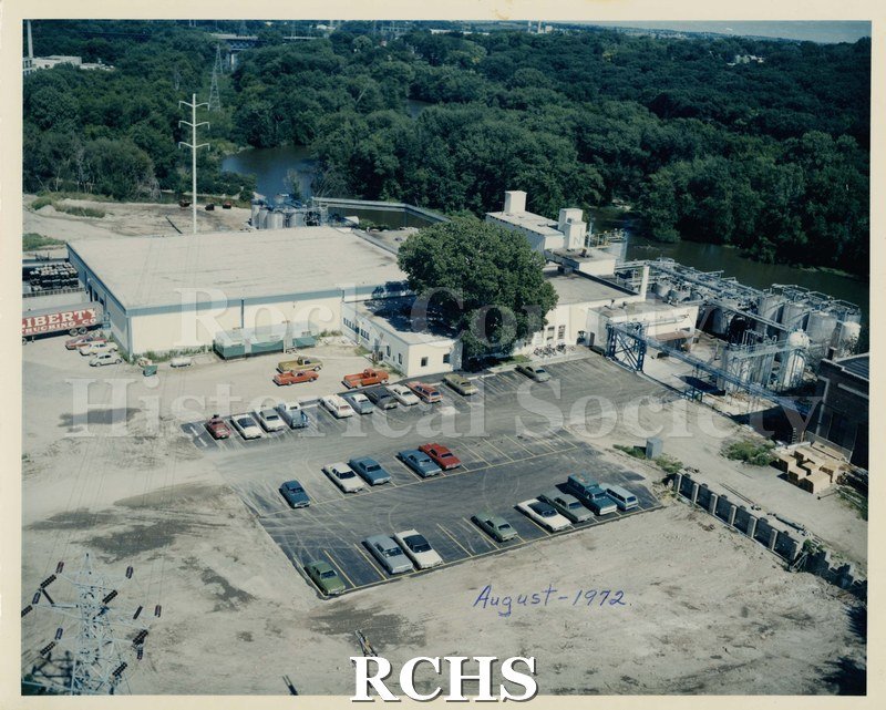 Colored photograph of Ashland Chemical, Janesville, WI. Shows a one-story factory with cars parked in the parking lot. There are oil storage tanks on the right side.