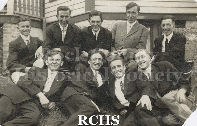 Black and white photograph of nine young men sitting in front of a building.