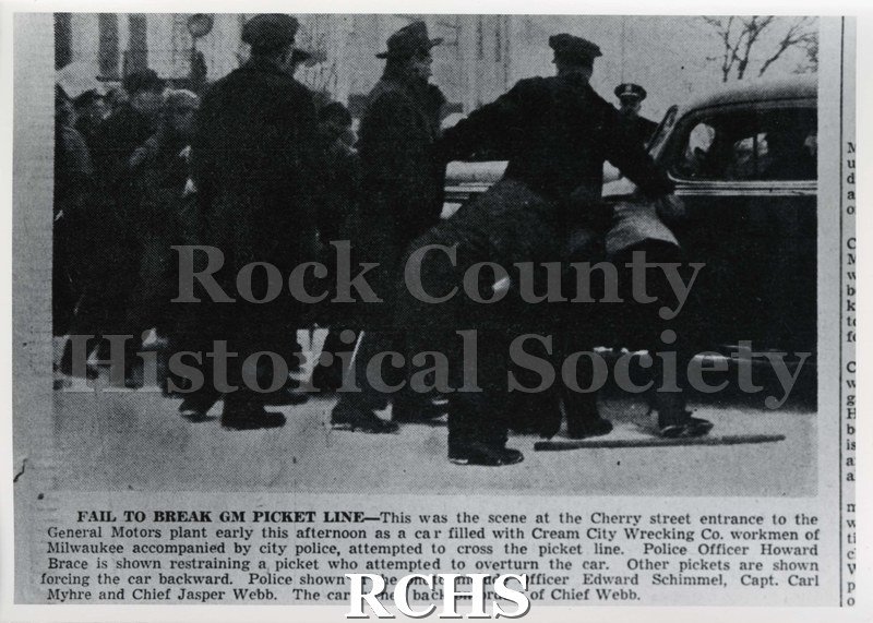 Black and white photograph of a newspaper article about a strike at GM. Picture shows police holding back cars trying to enter.