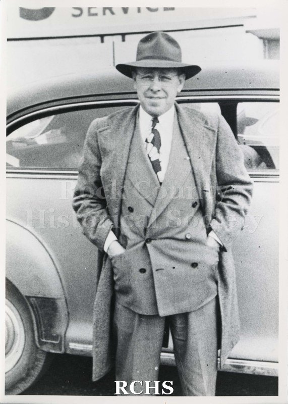 Black and white photograph of Gerald Litney. He is wearing a topcoat over a dark colored suit, white shirt, tie and hat. He is standing beside a car.