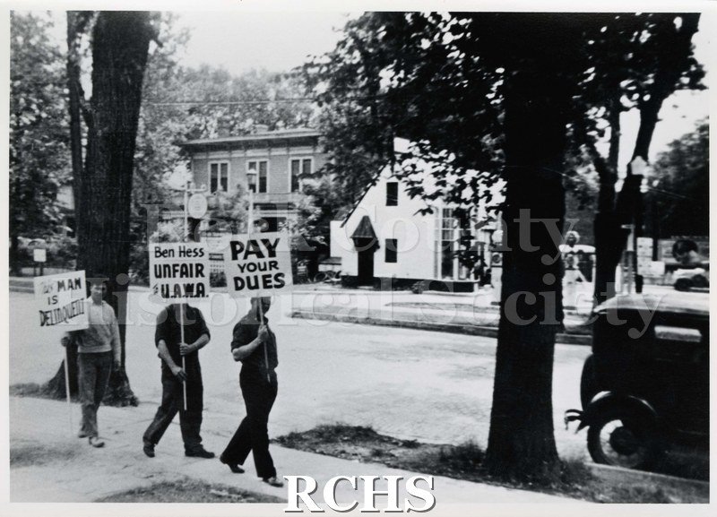 Black and white photograph of Ben Hess's house being picketed by UAWA 1977? Three men are on the sidewalk across the street from the house, bearing signs.