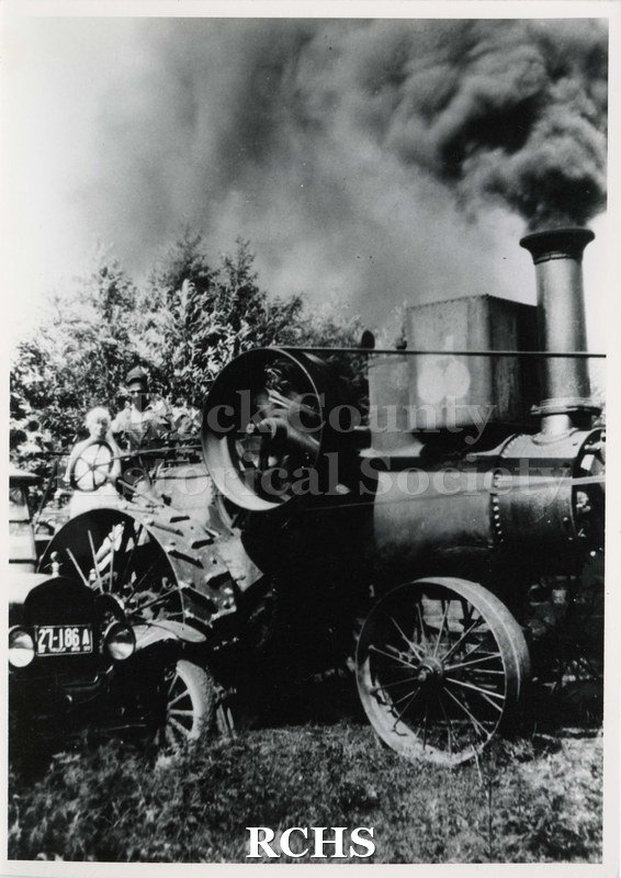 Black and white photograph of threshing oats near Afton. Shows a man and a woman on a threshing machine. License 27-186A is visible on car beside.