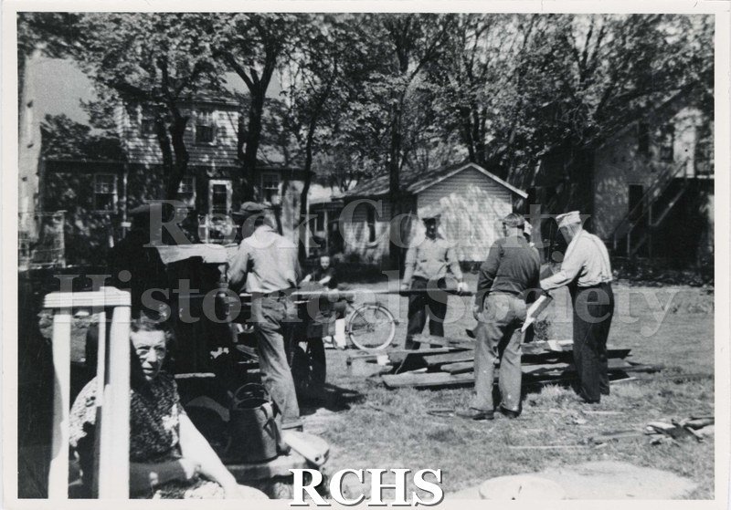 Black and white photograph of several men removing lumber from a building. There is a woman sitting on a porch in the left bottom corner.