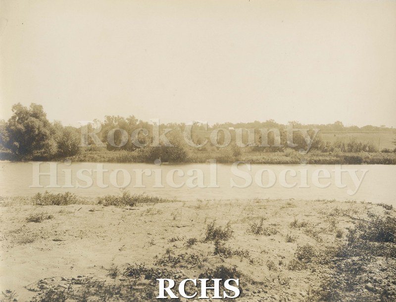 Black and white photograph of Rock River. Shows river in middle with grassy bank in front and trees in the background. A building is visible through the trees.