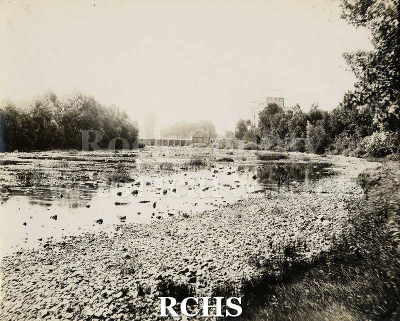 Black and white photograph of Rock River. Shows the rocks in front and the dam in the background.