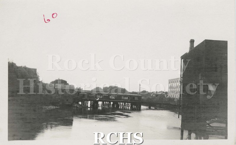 Black and white photograph of early Janesville. Shows the river in front with a pier extending into the river.