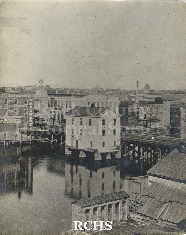 Black and white photograph of early Janesville. Shows a building in the river on pilings which is reflected in the river.