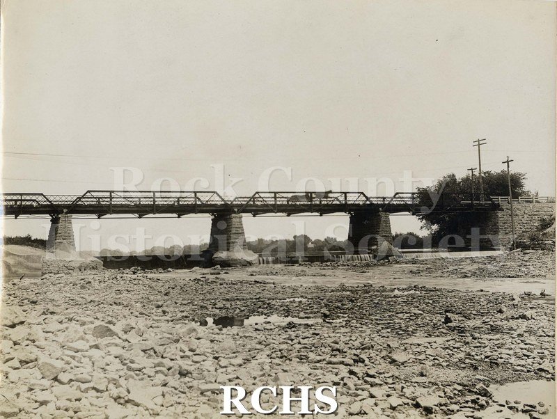Black and white photograph of the Rock River. Shows the bridge above the dam with a horse and wagon traveling along the bridge.