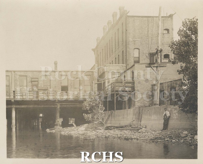 Black and white photograph of early Janesville, showing the river in front with a building on the right side.
