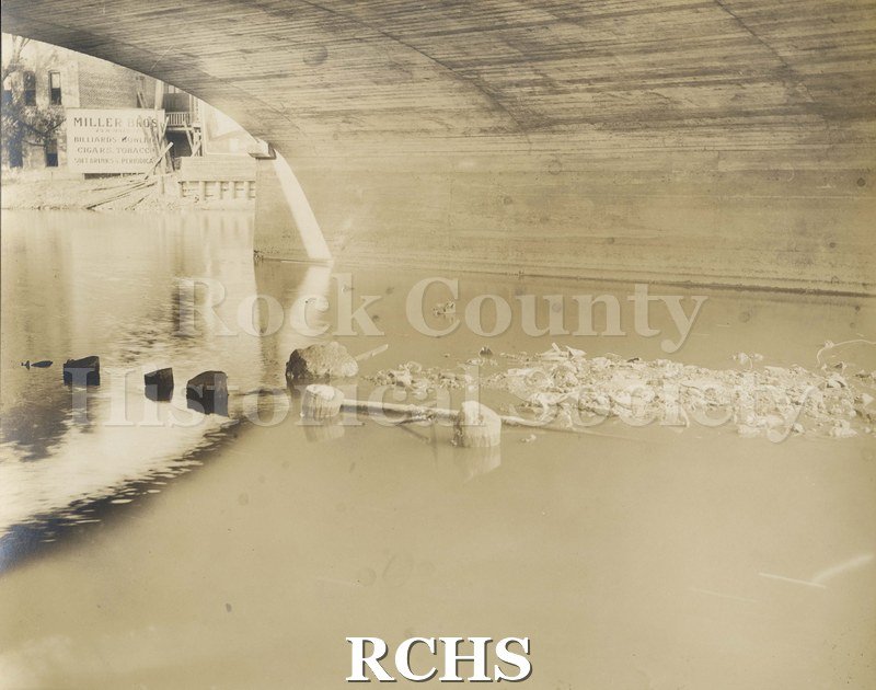 Black and white photograph of the inside of tunnel under Rock River. Shows water with rocks in the middle. To the left is a building with a sign: "Miller Bros".