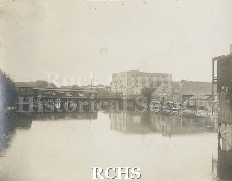 Black and white photograph of early Janesville. Shows the river in front with a pier extending into the river.