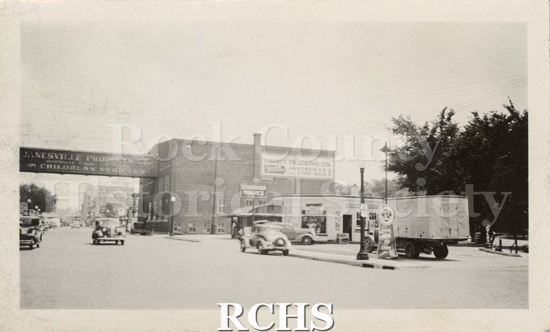 Black and white photograph of Janesville Products Company. Shows a large building with a sign over the street: Janesville Products Co.