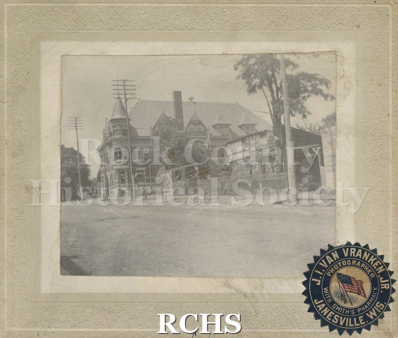 Black and white photograph of the old YMCA. Unusual unidentified shack-like building in the foreground of the photo. On bottom is a seal: "J. I. Van Vranken, Jr.