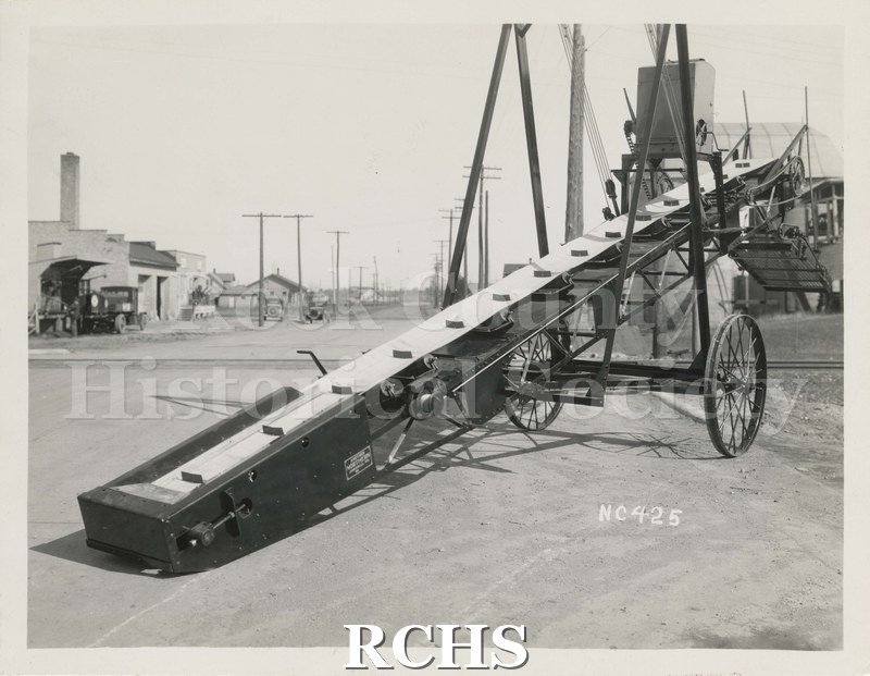 Black and white photograph of a Northern Conveyor & Mfg. Co. conveyor on the street. On the conveyor is a sign: ""Another Northern Janesville Wis".