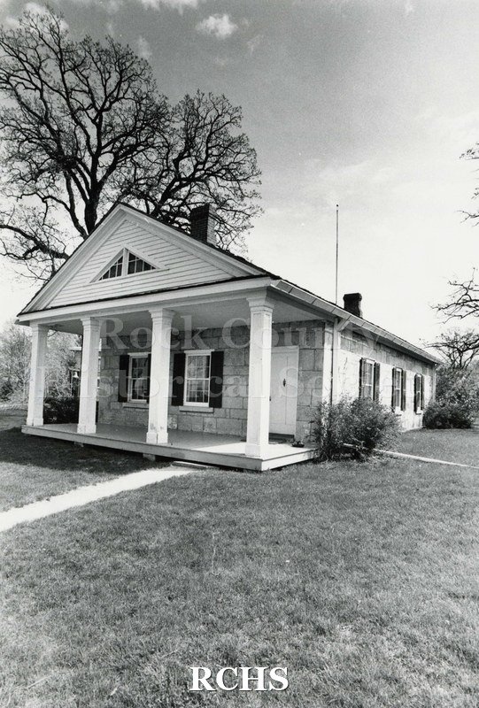 Black and white photograph of the Stone House. Shows the pillared porch, three windows on the right side and trees in the background.