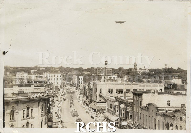 Black and white photograph of military parade on Milwaukee Street c. 1942. Towers of Rock County Courthouse and Jefferson School visible.
