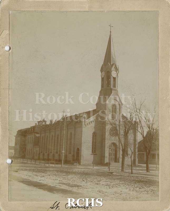 Black and white photograph of St. Patrick Church, Janesville, WI. Shows an early church building with a clock tower. On bottom: "St. Patrick".