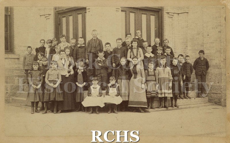 Sepia photo of Washington School, Janesville, WI c. 1890. Shows a group of school children and teachers standing in front of a school building.