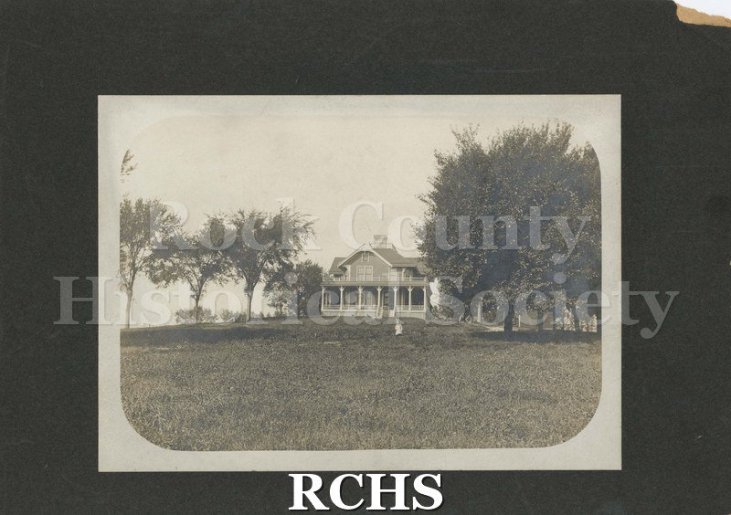 Black and white photograph of the Woodruff Farmhouse, Janesville, WI. Shows a farmhouse with a pillared porch and a balcony on top. House is surrounded by trees.