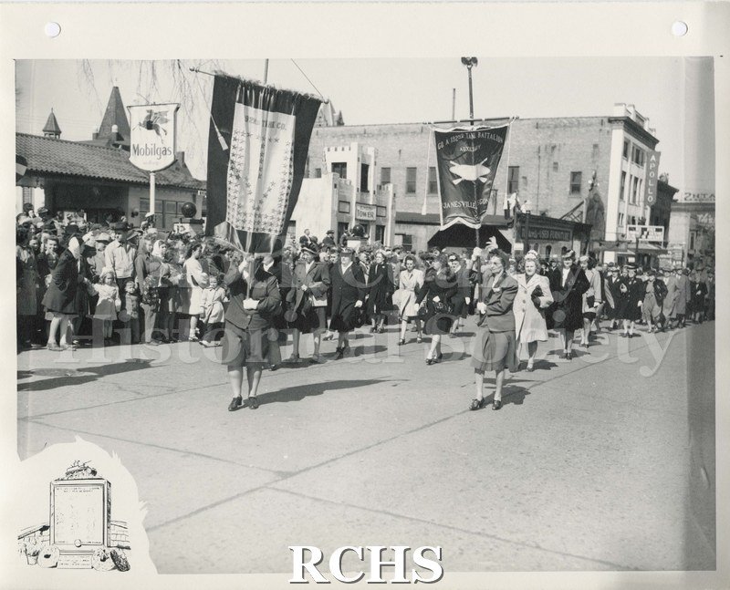 Black and white photograph of a military parade through downtown Janesville. Women are marching in front of the Mobilgas station and Apollo Theatre.