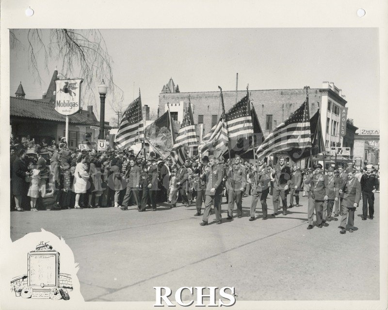 Black and white photograph of a military parade through downtown Janesville. Men in front are bearing American flags.