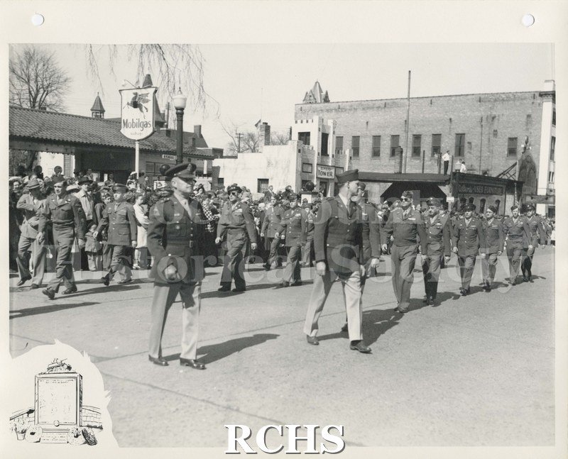 Black and white photograph of a military parade through downtown Janesville. Men are turning the corner of the parade route.