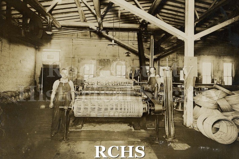 Black and white photograph of Harris Factory, Janesville, WI? There are two men standing by a machine that is winding wire.