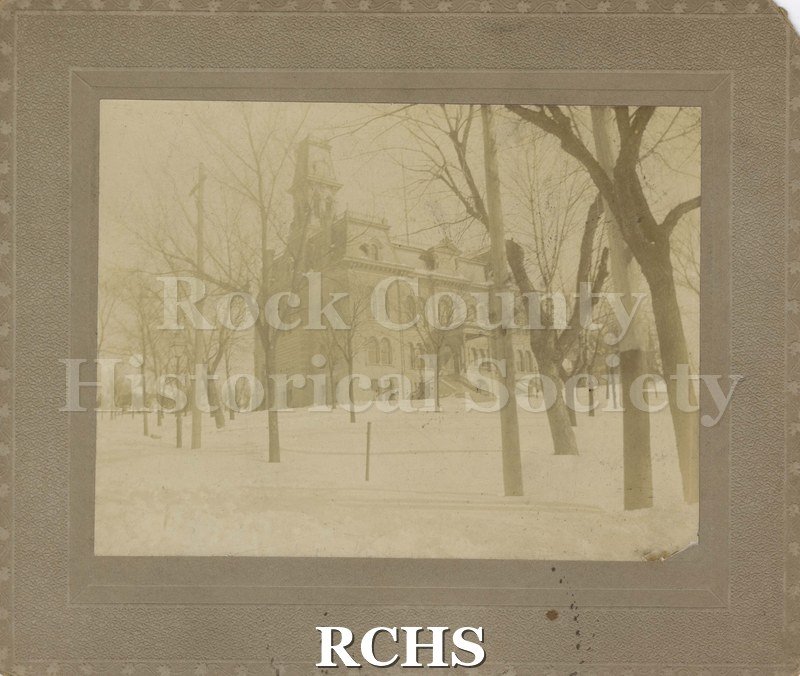 Black and white photograph of Janesville Court House. Building 2 1/2 stories with mansard roof and clock tower. Foreground is rows of trees and snow covered ground.