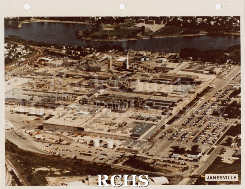 Colored photograph of General Motors from the air. Rock River visible in background. Buildings surrounded by streets and parked cars.
