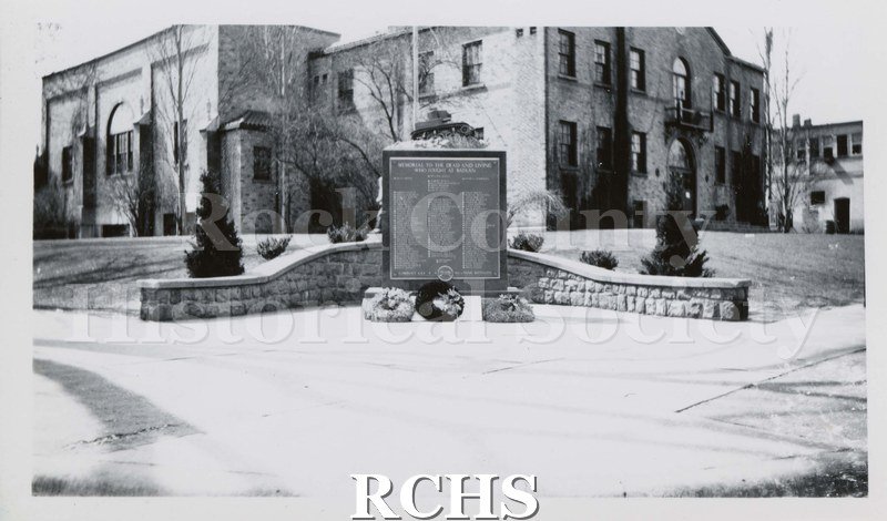 Black and white photograph of Janesville Armory from corner with sign at center. Road and sidewalk to curved stone wall and floral arrangements highlight sign.