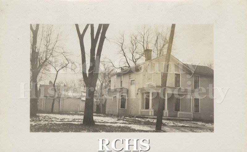 Black and white photograph of Henry Bull house and garage. Two story with light colored siding and white trim. Snow on ground, Trees in foreground and background.
