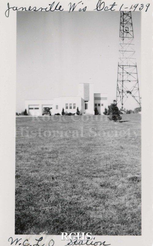 Black and white photograph of WCLO Station 1939 showing white building in distant background with metal tower to right. Car parked in front of garage.