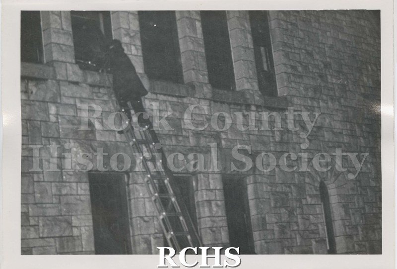 Black and white photograph of fire at Beloit College Eaton Chapel. Shows fireman on ladder reaching into window on second story of stone builidng.