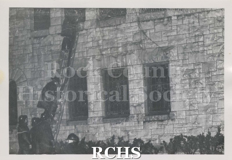 Black and white photograph of the fire at Beloit College Eaton Chapel. Shows two firemen on ladder to window on second story of stone builidng.