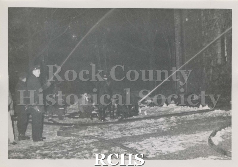 Black and white photograph of the fire at Beloit College Eaton Chapel. Shows several firemen standing or kneeling holding hoses with streams of water directed up.
