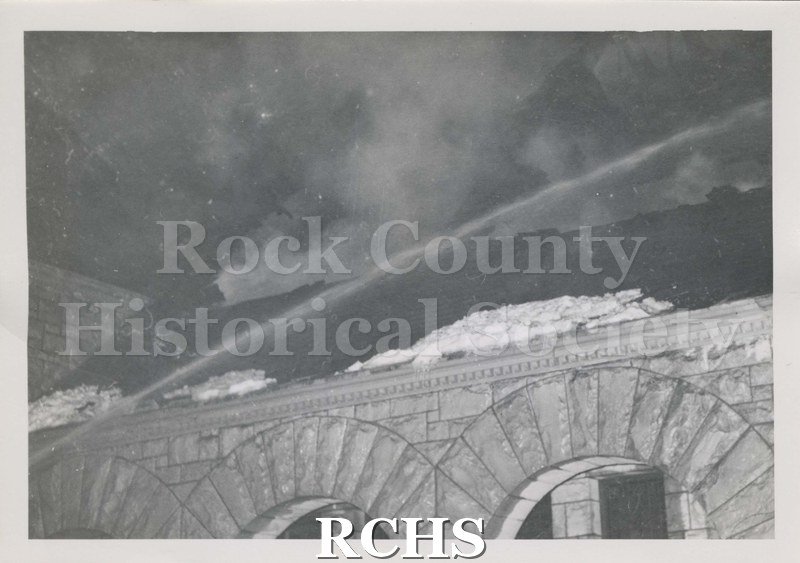 Black and white photograph of the fire at Beloit College Eaton Chapel. Shows part of stone building with arches in foreground, snow on roof, stream of water, smoke.