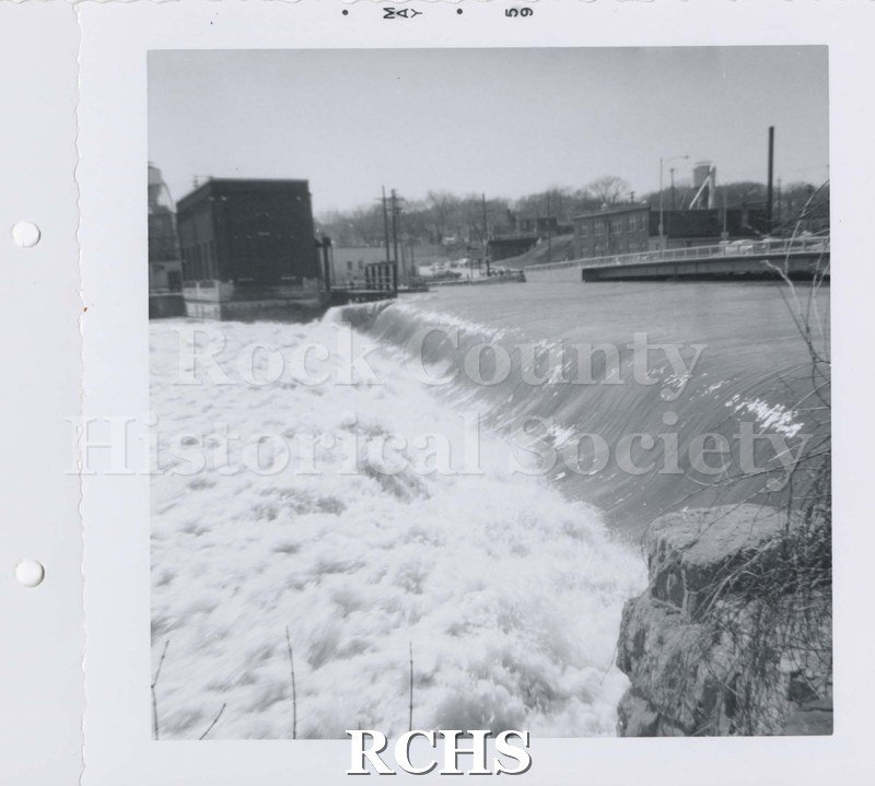 Black and white photograph of Centerway Dam. Shows turbulant water flowing over Centerway Dam with bridge on left. Square building to left of photo.